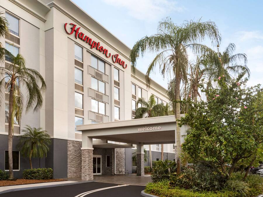 A Hampton Inn hotel with a modern facade, a welcoming entrance, and a landscaped outdoor area with palm trees and shrubs.