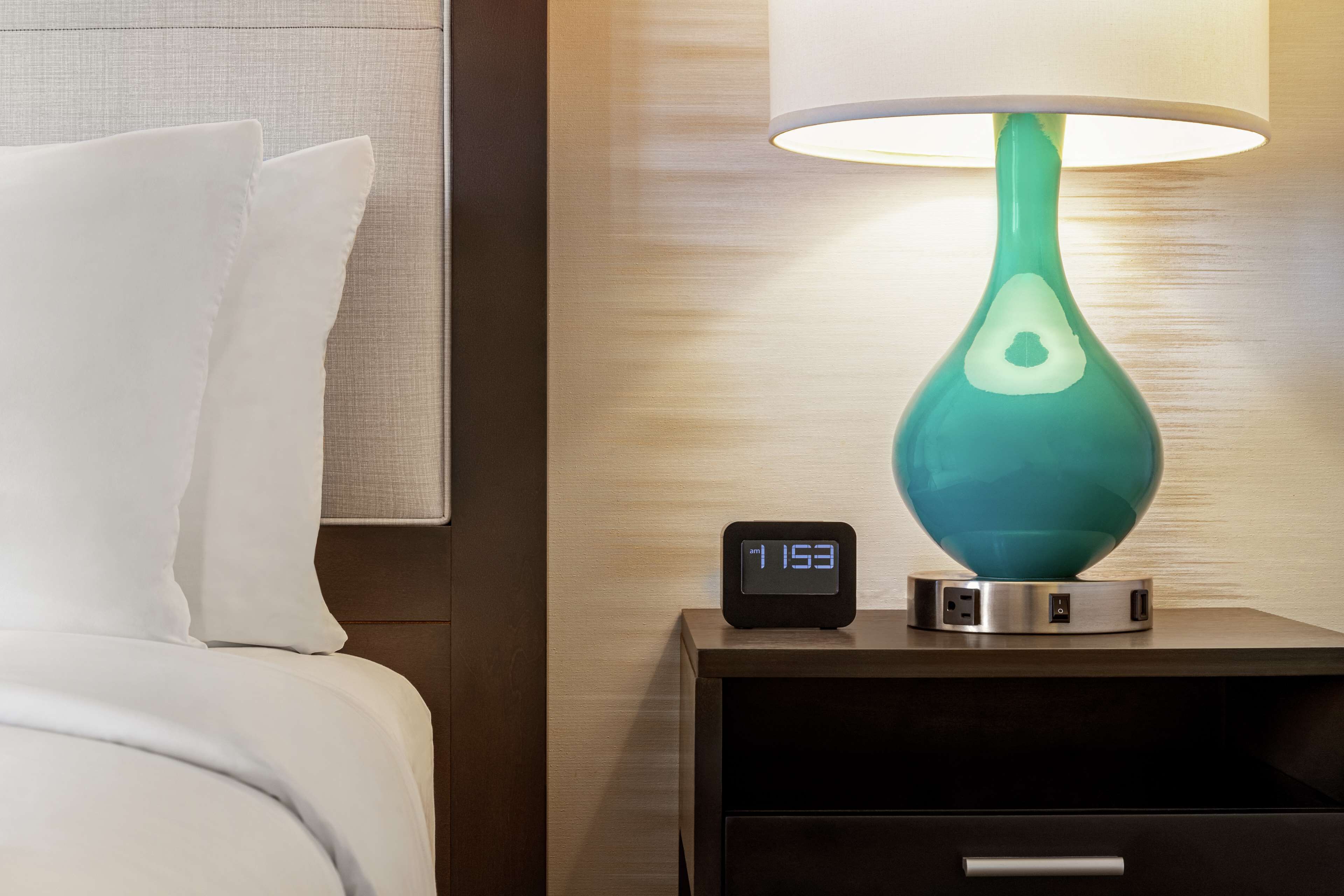 A neatly made bed with white linens, a teal-colored lamp on a bedside table, and a digital clock displaying the time.