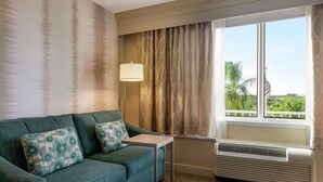 A room with a green sofa, patterned pillows, a small table, a lamp, and a window with an air conditioner. - Hampton Inn Ft. Lauderdale-West/Pembroke Pines (Pembroke Pines)