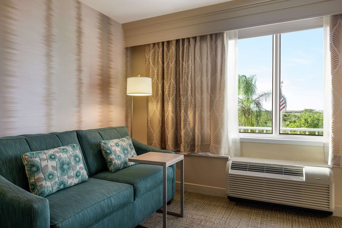 A room with a green sofa, patterned pillows, a small table, a lamp, and a window with an air conditioner.