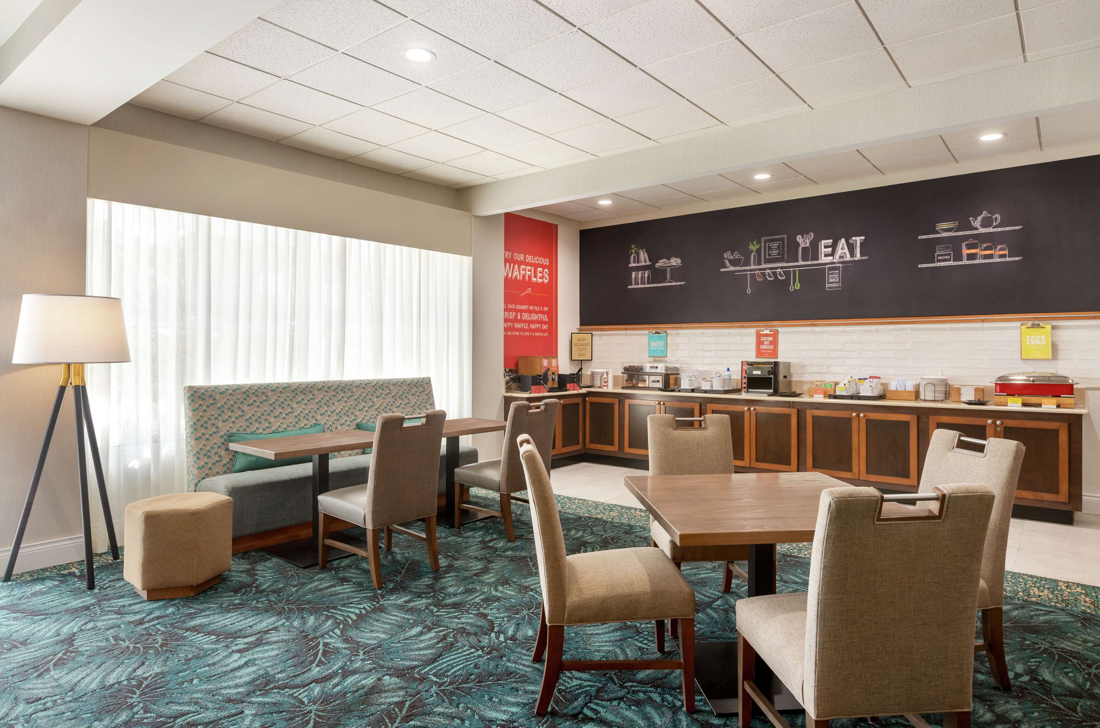 A hotel room with a dining area, kitchenette, and a large window with blinds.