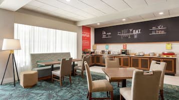 A hotel room with a dining area, kitchenette, and a large window with blinds.