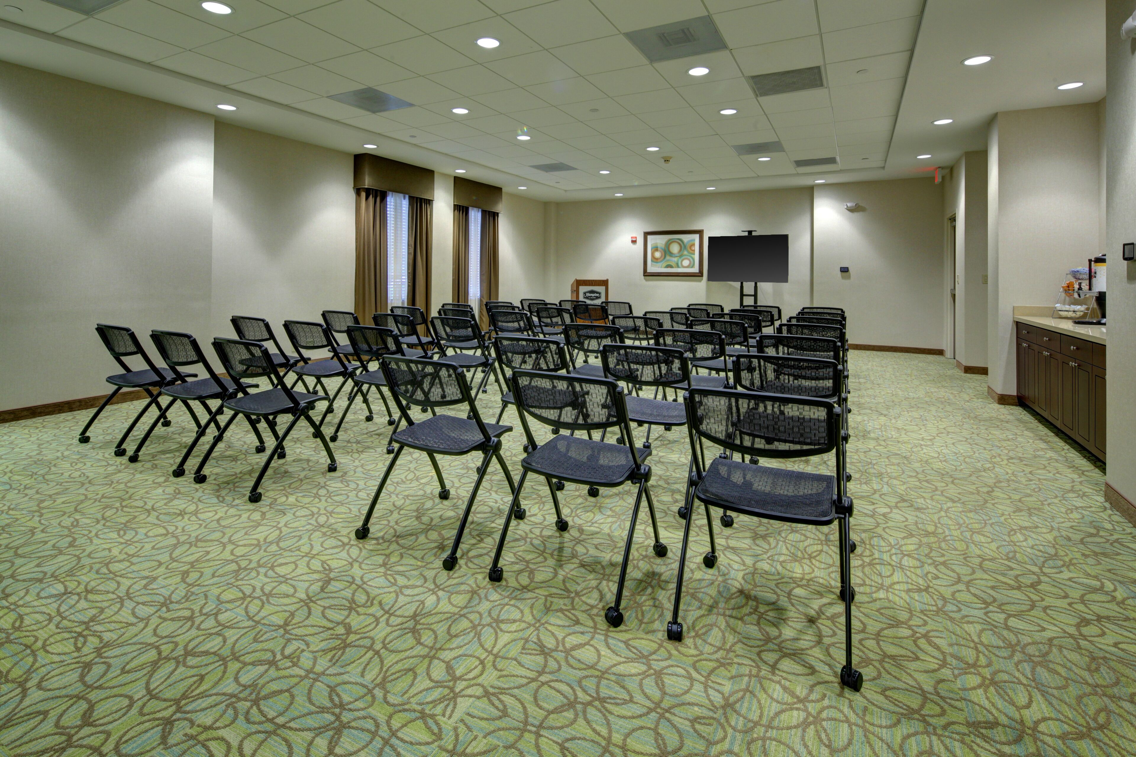 A conference room with rows of foldable chairs, a projector screen, and a framed picture on the wall.