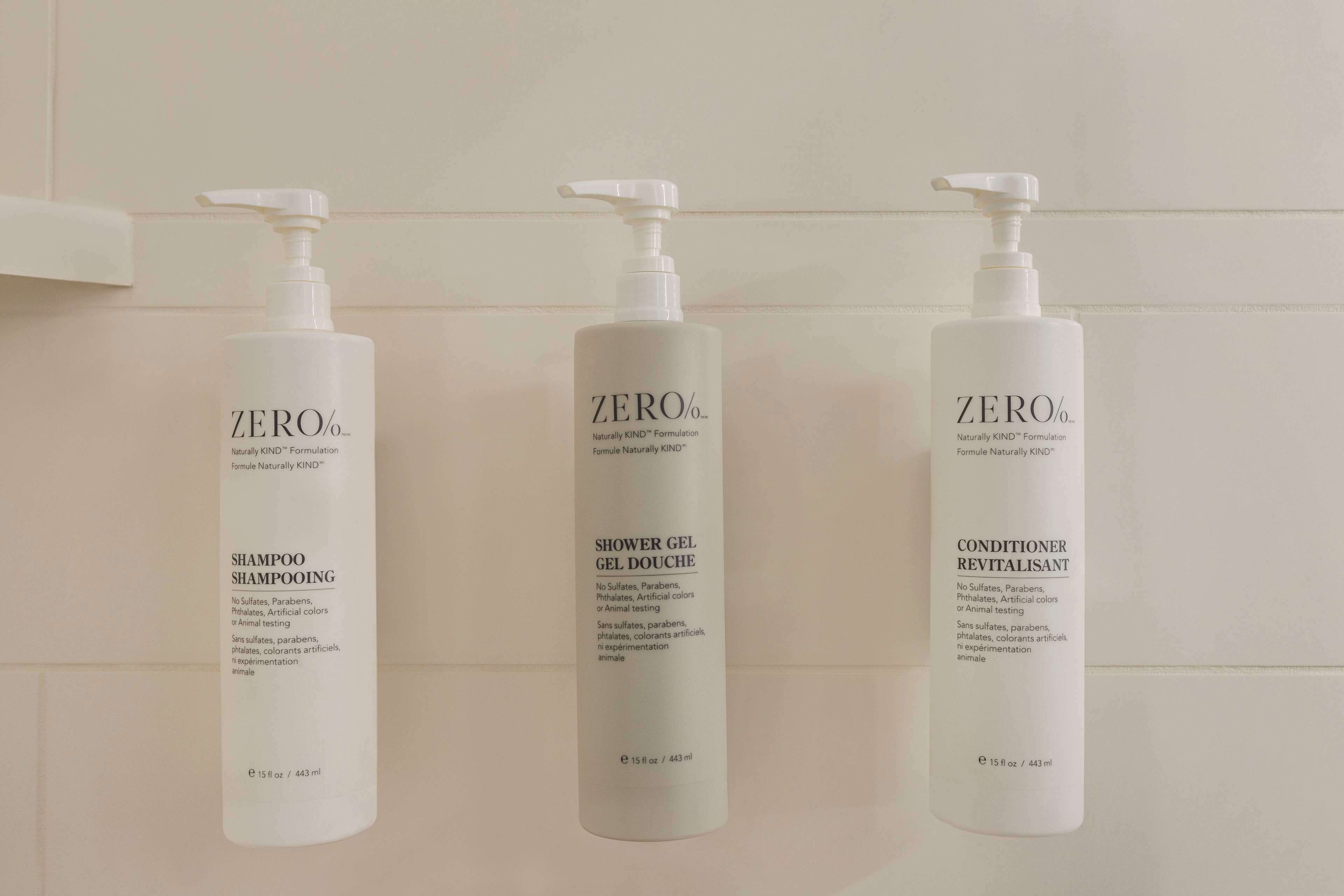 Three bottles of Zero% shampoo, shower gel, and conditioner on a white tiled wall.