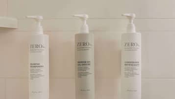 Three bottles of Zero% shampoo, shower gel, and conditioner on a white tiled wall.