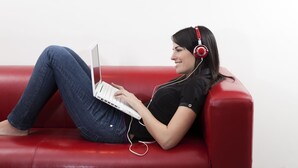 Een persoon zit op een rode leren bank en gebruikt een laptop met koptelefoon.