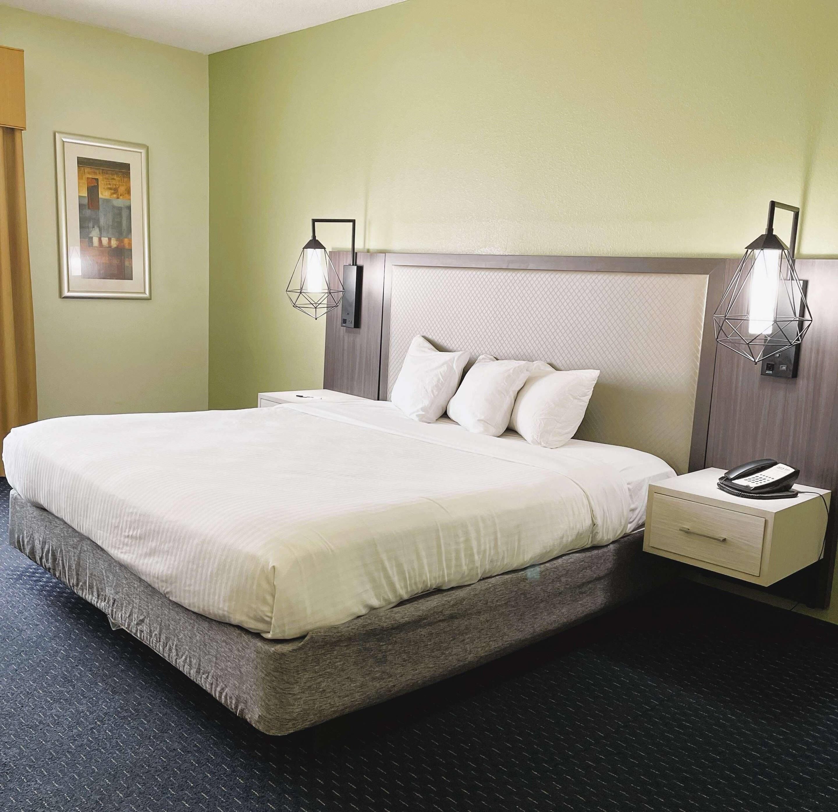 Quarto de hotel com uma cama grande, duas mesas de cabeceira, um criado-mudo com telefone e luminárias fixadas na parede.