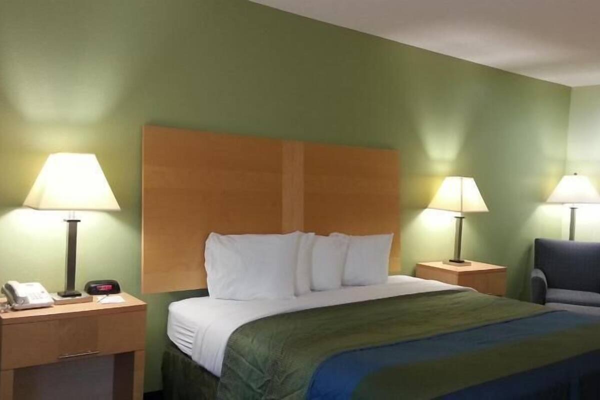 A hotel room with a bed, two bedside lamps, a chair, a desk with a phone, and a nightstand.