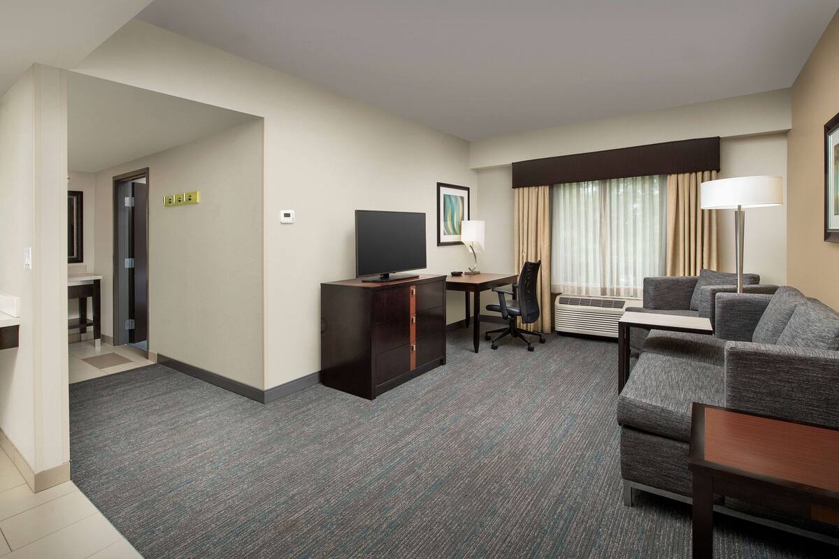 A hotel room with a grey carpet, a sofa, a TV, a desk, and a lamp.