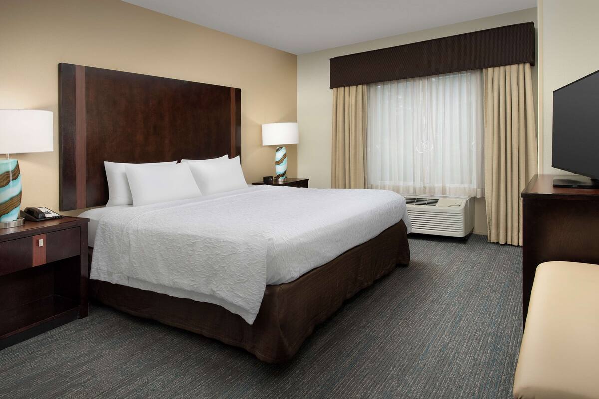 A hotel room with a large bed, a bedside table with a lamp, a TV, and a window with curtains.