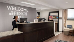 A hotel reception area with a dark wood reception desk, a large welcome sign, a computer, and a small shop in the background. - Hampton Inn & Suites Alpharetta (Alpharetta)