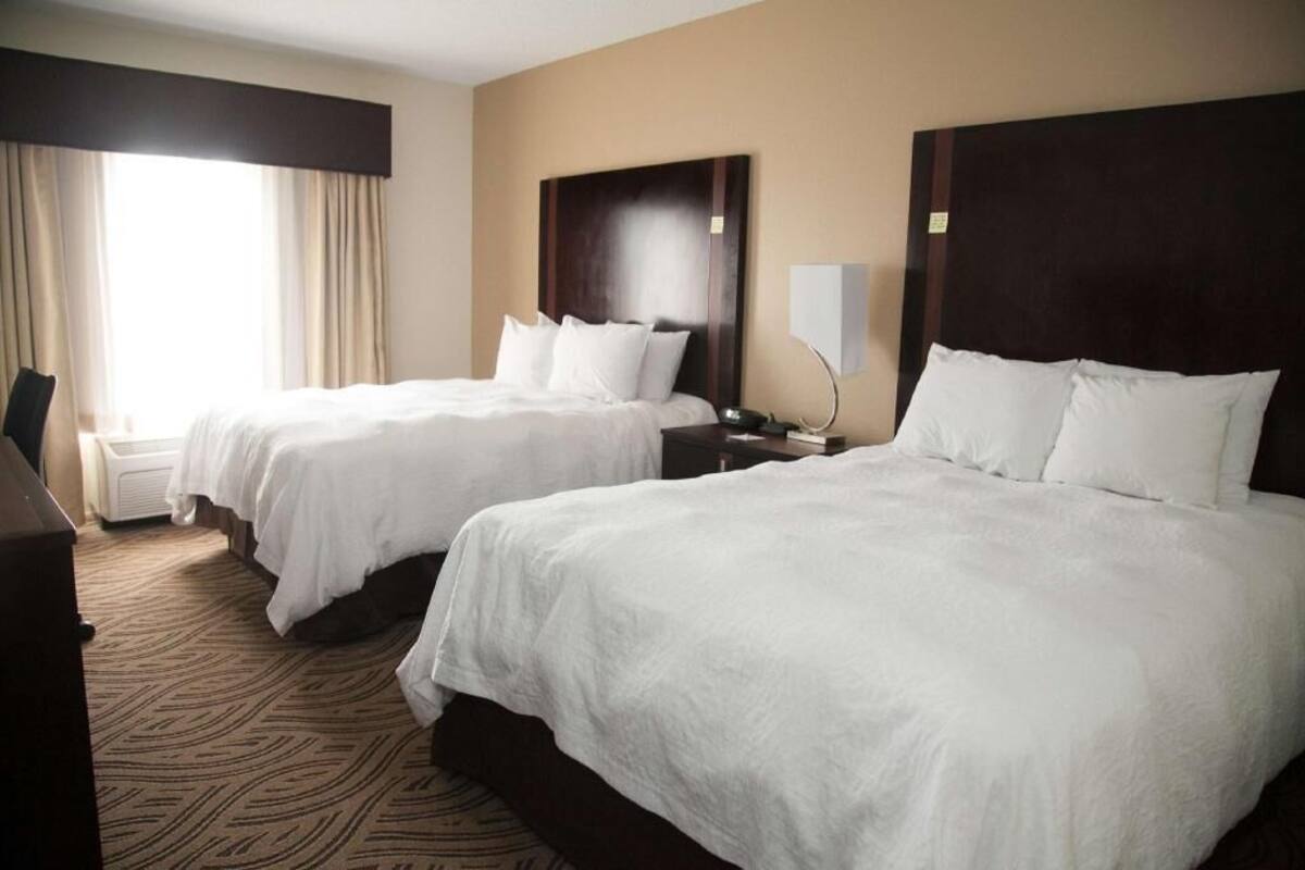 A hotel room with two beds, a desk, a chair, a lamp, and a window with curtains.