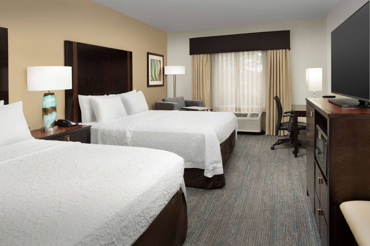 A hotel room with two beds, a desk, a chair, a TV, and a window with curtains.