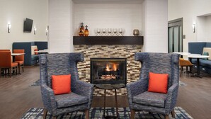 A modern lounge area with a fireplace, two armchairs with orange cushions, and a patterned rug. - Hampton Inn & Suites Alpharetta (Alpharetta)