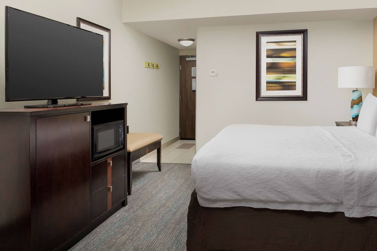 A hotel room with a bed, a TV, a desk with a chair, a lamp, and a painting on the wall.