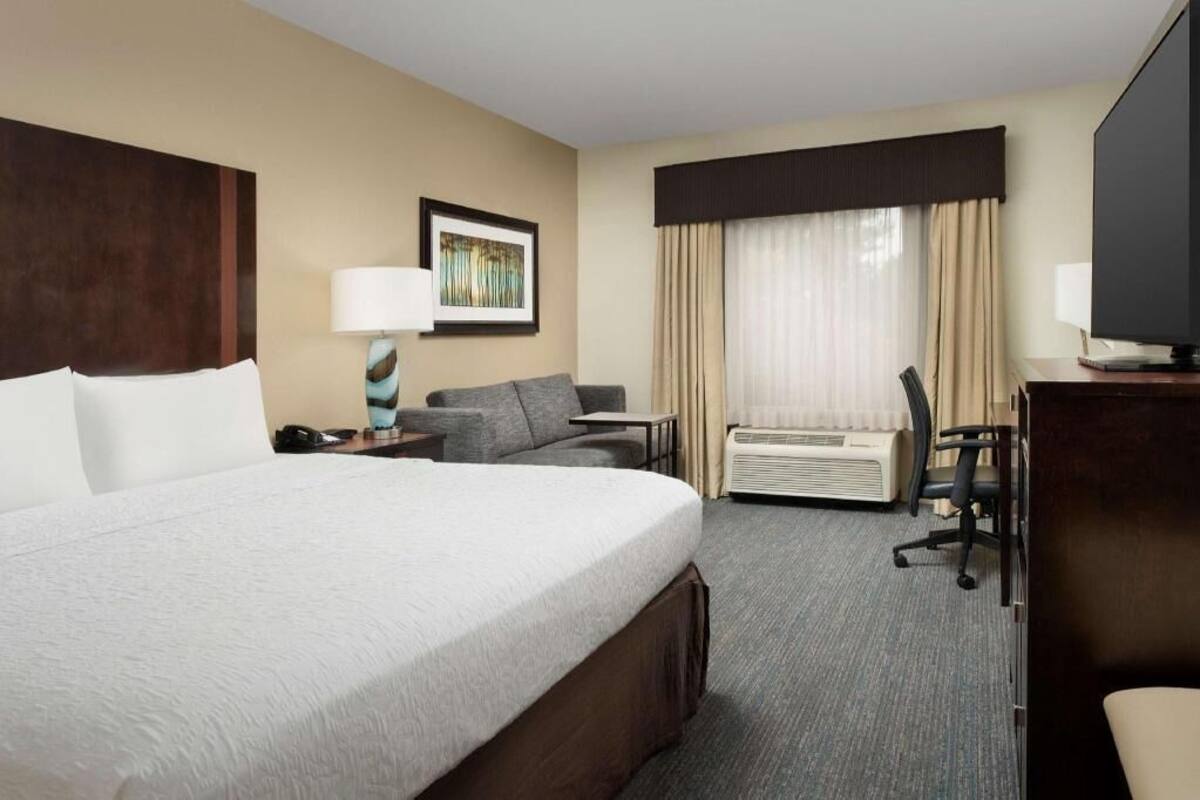A hotel room with a large bed, a sofa, a desk with a chair, a television, and an air conditioning unit.