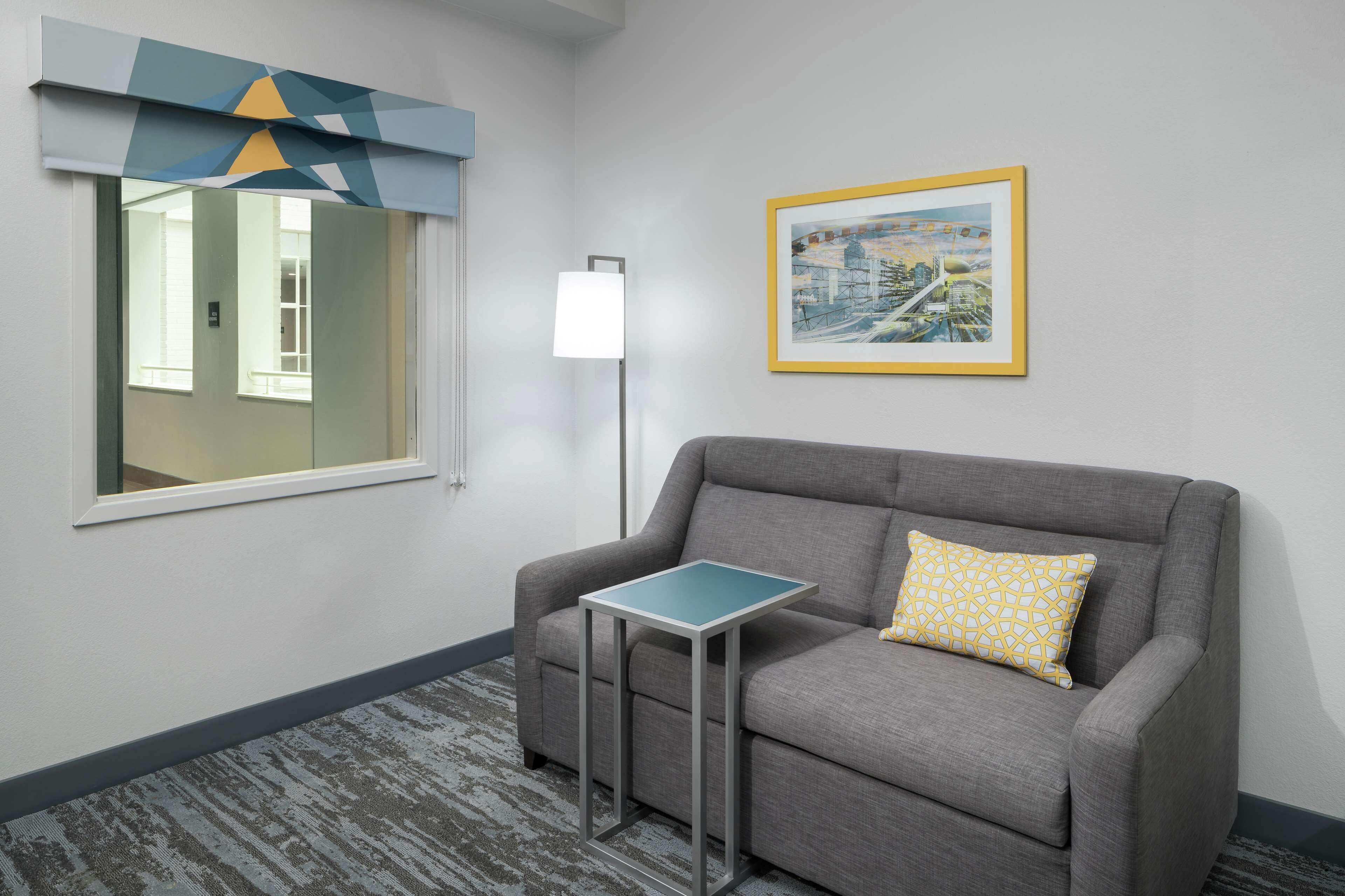 A modern hotel room with a grey sofa, a small table, a framed cityscape painting, and a window with a geometric curtain.