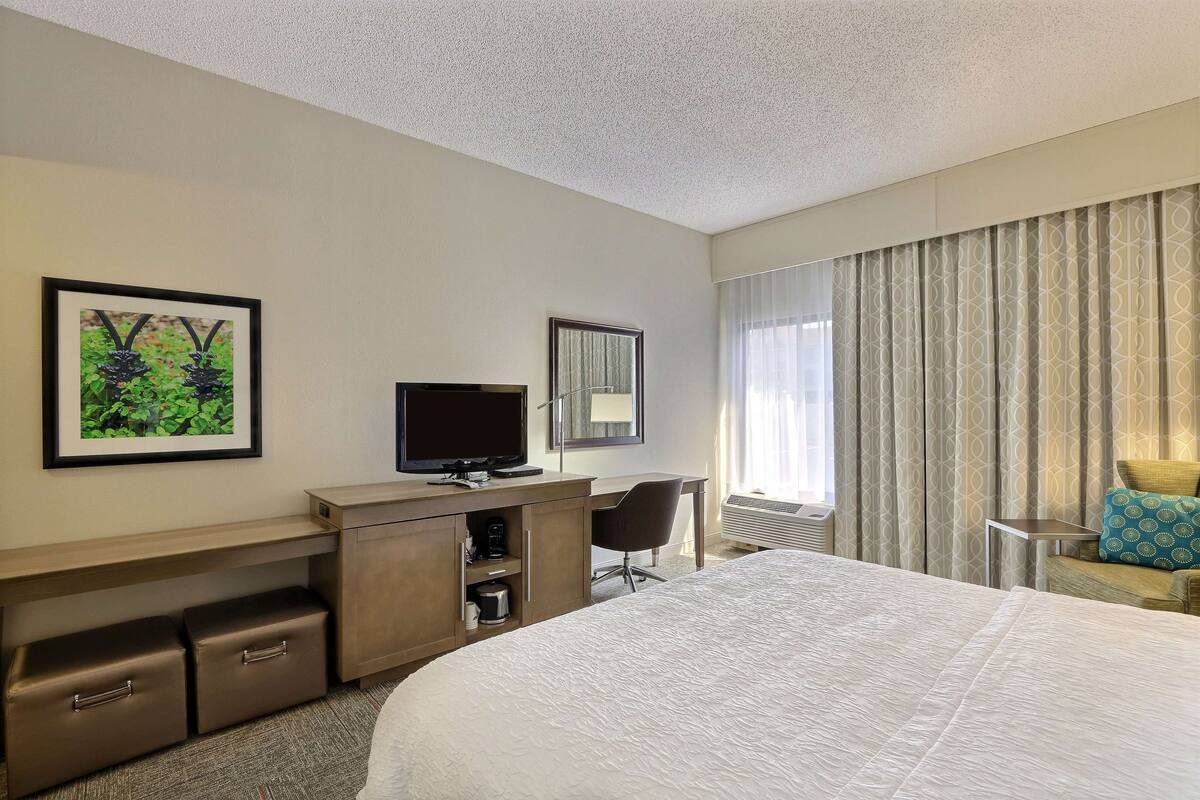 Ein Hotelzimmer mit einem großen Bett, einem Schreibtisch mit Fernseher, einem Sessel und einem Fenster mit Vorhängen.