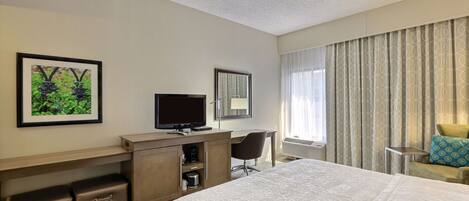 Habitación de hotel con una cama grande, un escritorio con televisor, una silla y una ventana con cortinas.