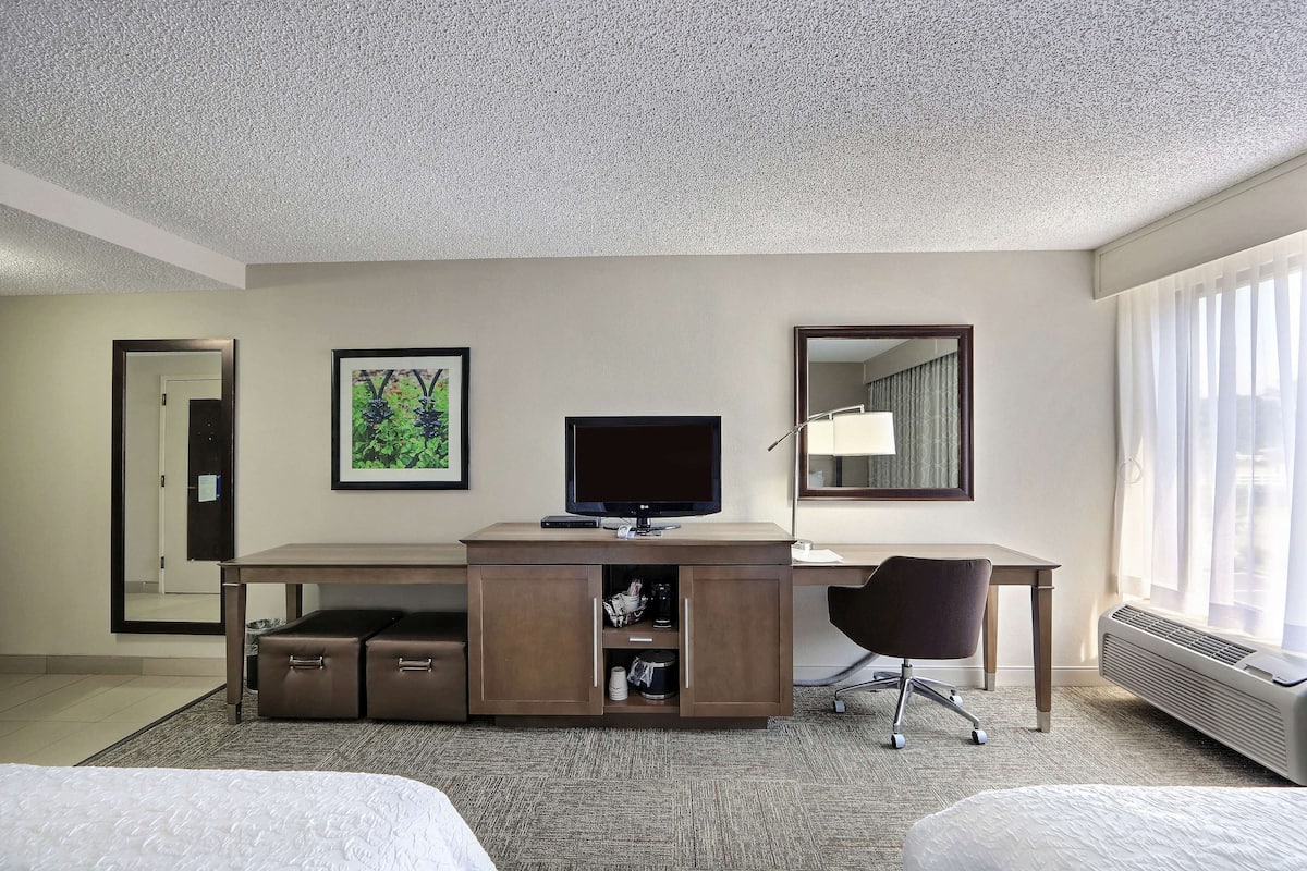 Ein Hotelzimmer mit Bett, Schreibtisch mit Computer, Stuhl, Spiegel und einem Fenster mit Vorhängen.