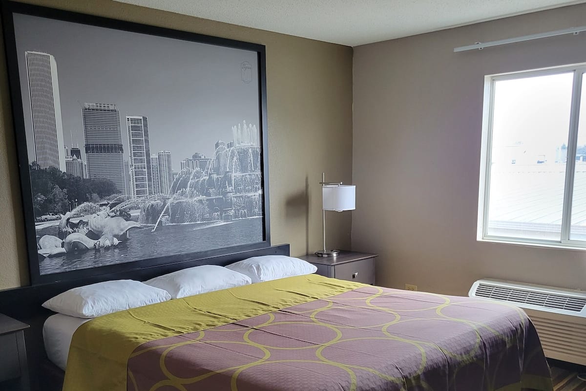 A hotel room with a large bed, a cityscape artwork on the wall, a bedside table with a lamp, and a window with a view.