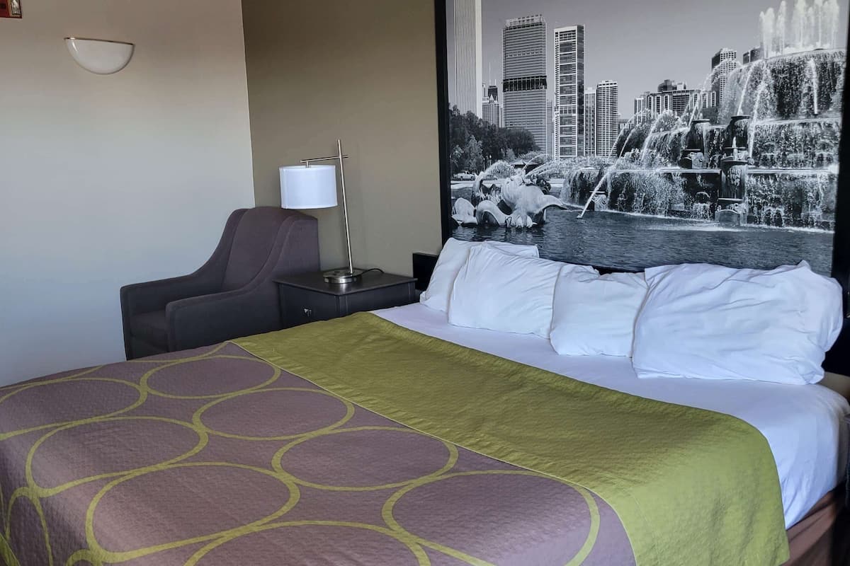 A hotel room with a bed, a chair, a nightstand, a lamp, and a large window with a cityscape view.