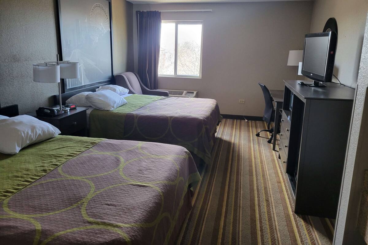 Ein Hotelzimmer mit zwei Betten, einem Schreibtisch mit Computer, einem Fernseher und einem Fenster mit Vorhängen.