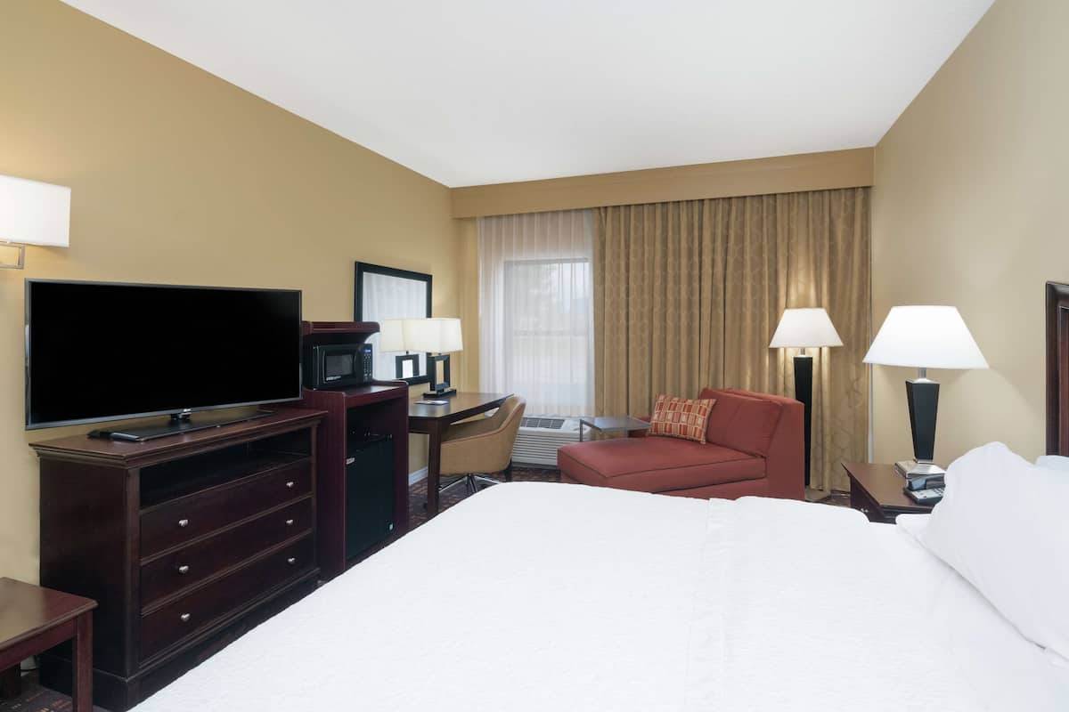 Quarto de hotel com uma cama grande, uma televisão de tela plana, uma escrivaninha com cadeira, uma poltrona vermelha e duas luminárias de cabeceira.
