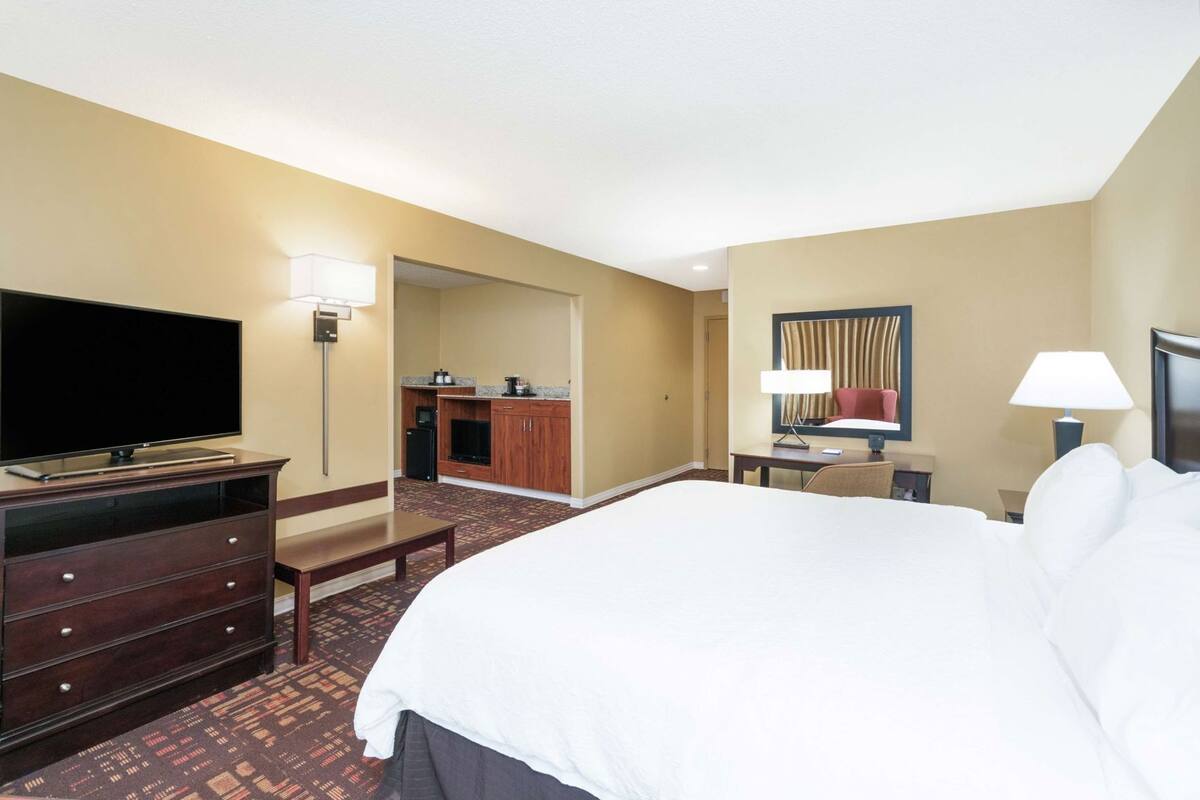 Ein Hotelzimmer mit einem großen Bett, einem Fernseher, einem Schreibtisch mit Lampe, einem Stuhl und einer Küchenzeile.