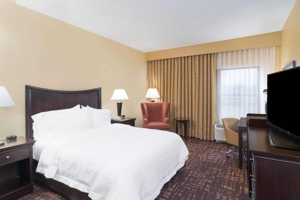 Quarto de hotel com uma cama grande, duas luminárias de cabeceira, uma cadeira, uma escrivaninha com televisão e uma janela com cortinas.