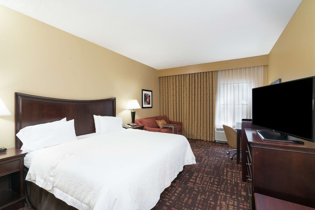 Quarto de hotel com uma cama grande, uma escrivaninha com televisão, uma cadeira, um abajur e uma janela com cortinas.