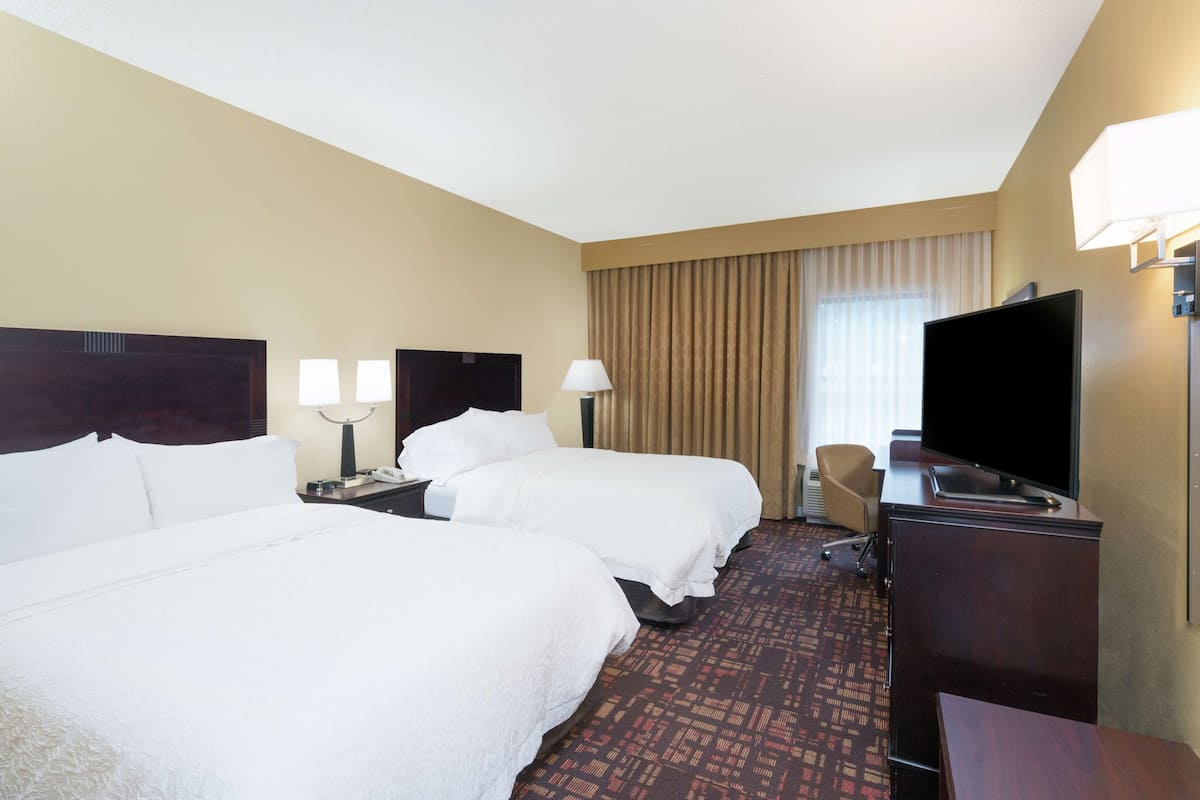 Quarto de hotel com duas camas, uma escrivaninha, uma cadeira, uma televisão e um abajur.