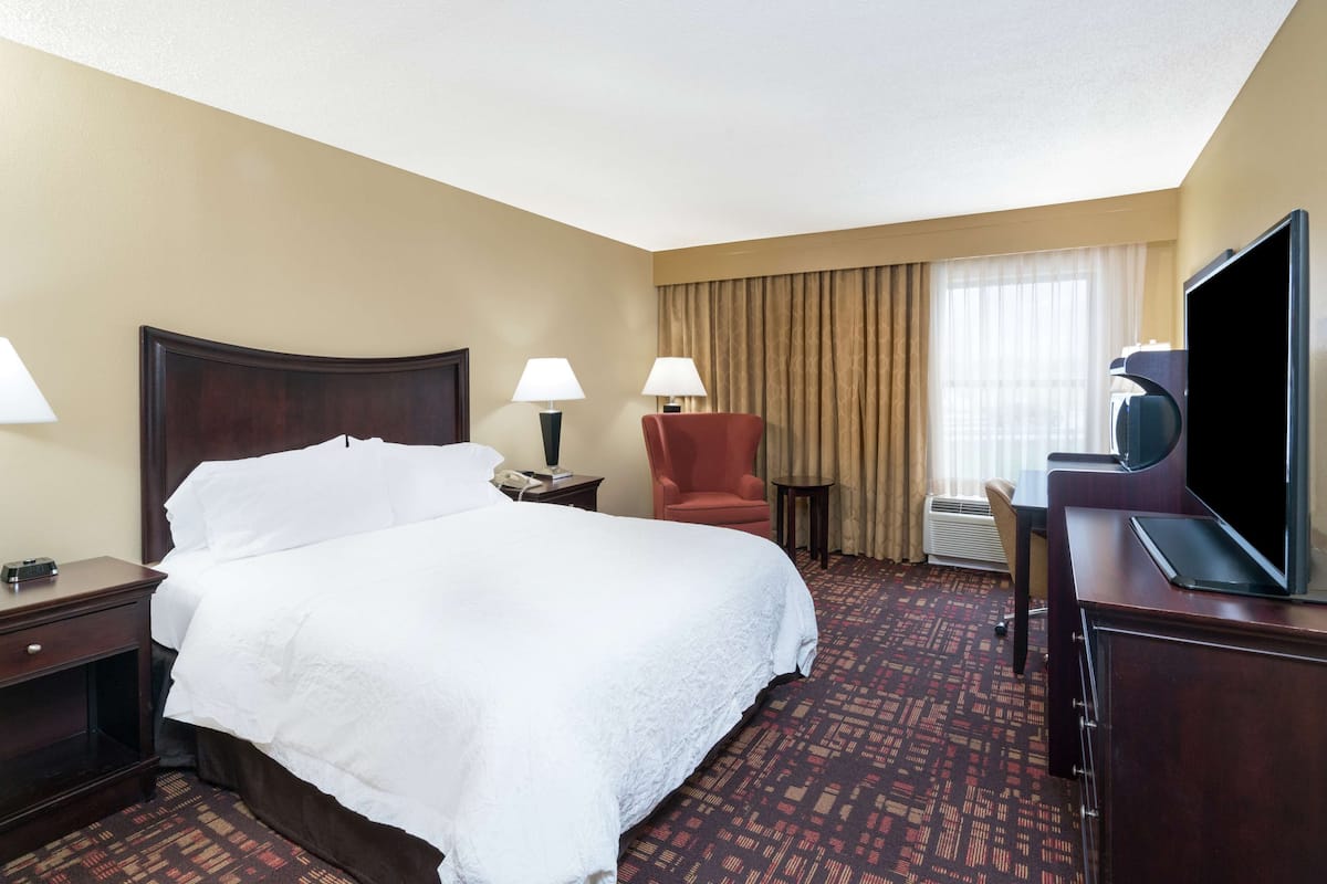 Quarto de hotel com uma cama grande, duas luminárias de cabeceira, uma poltrona vermelha, uma televisão e uma escrivaninha com um computador.