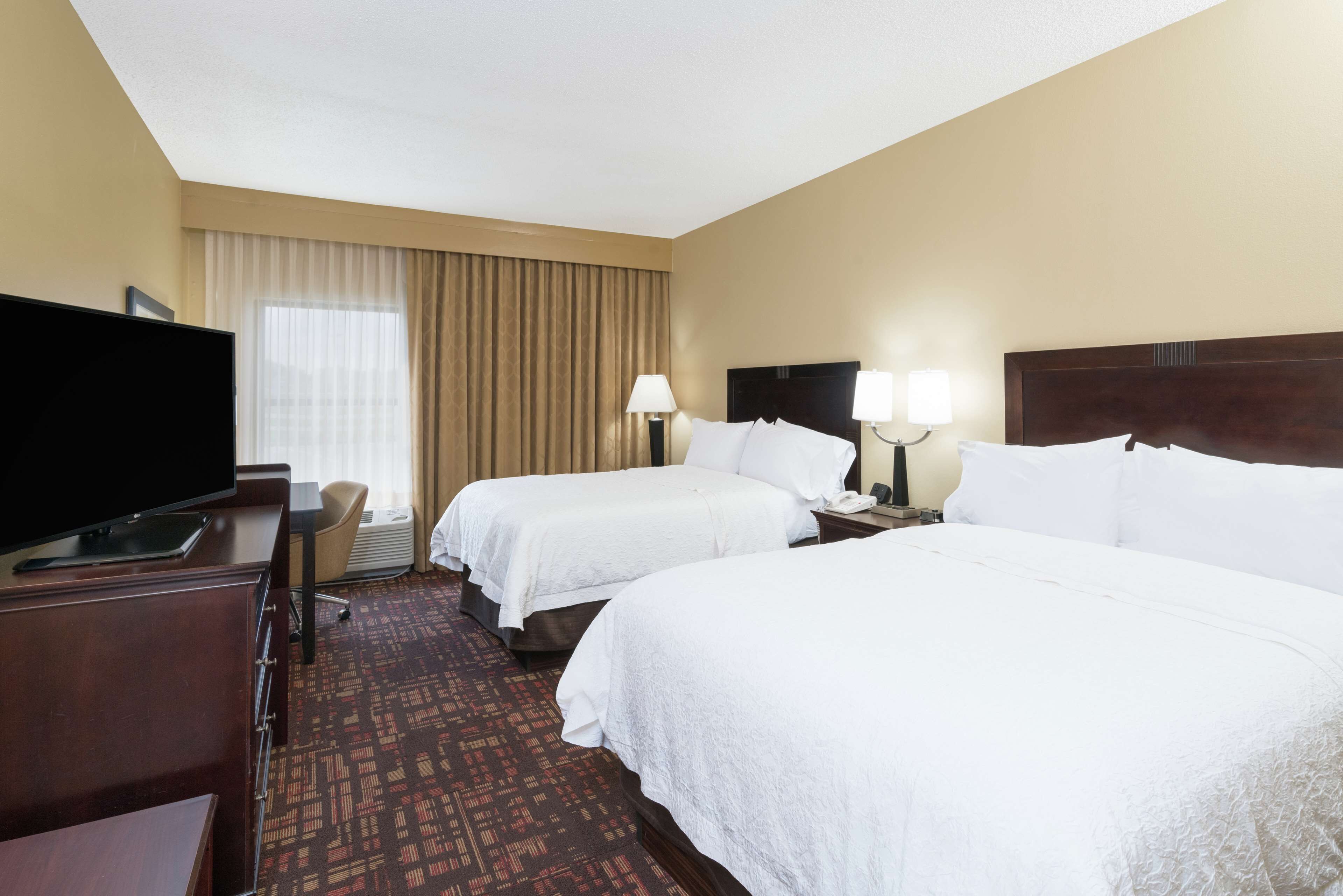 A hotel room with two beds, a TV, a desk, and a window with curtains.