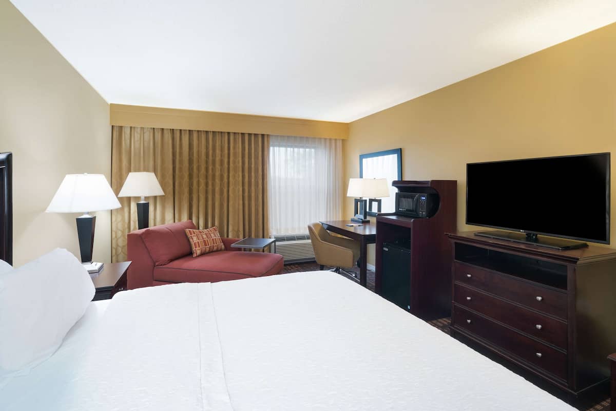 Quarto de hotel com uma cama grande, uma escrivaninha com computador, uma televisão e duas luminárias.