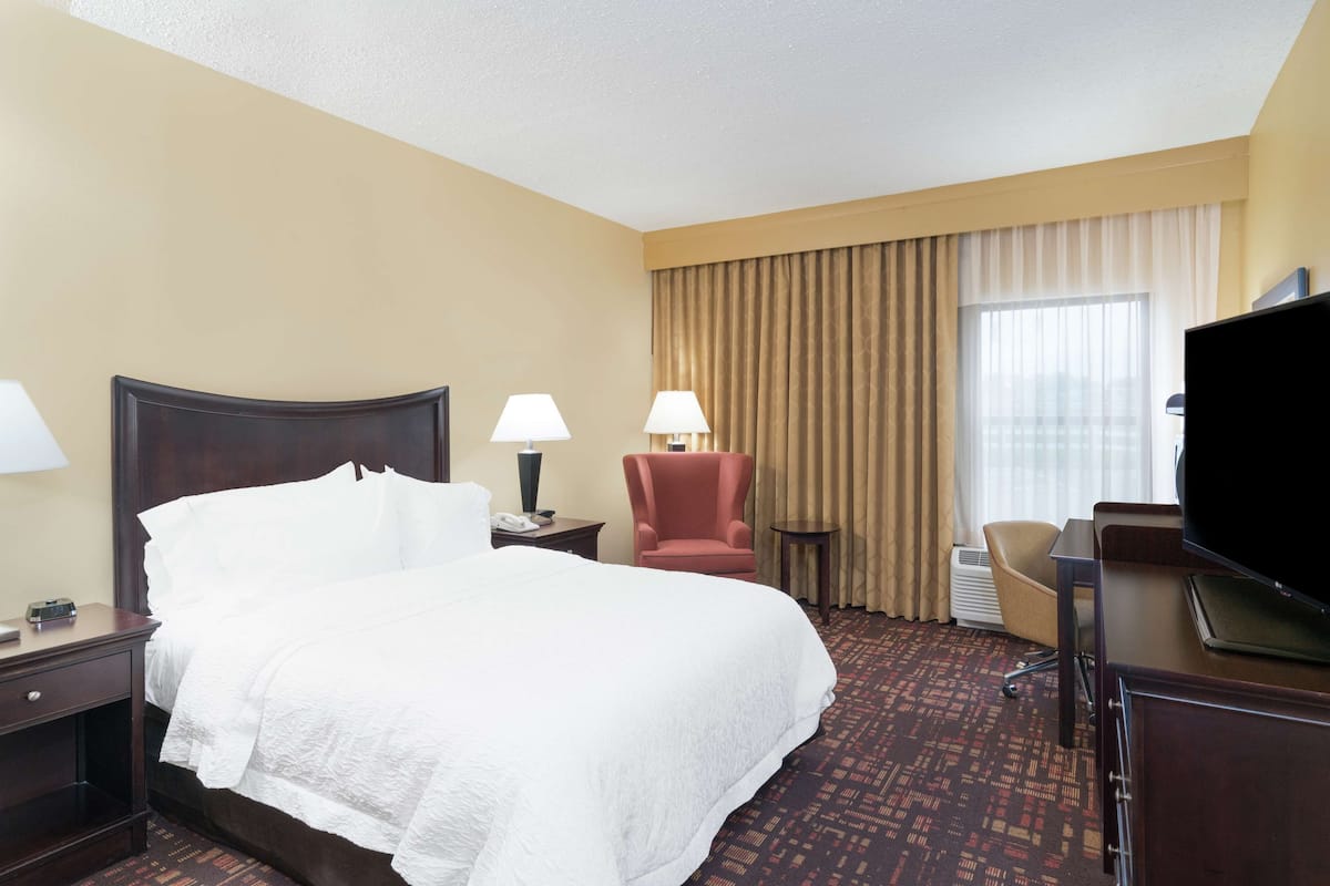 Quarto de hotel com uma cama grande, duas luminárias de cabeceira, uma poltrona vermelha, uma escrivaninha com televisão e uma janela com cortinas.