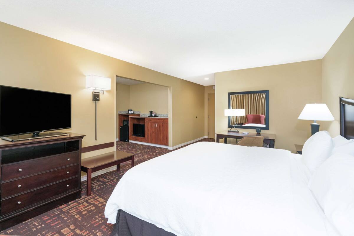 Quarto de hotel com uma cama grande, televisão, escrivaninha com cadeira, abajur e banheiro visível através de uma porta aberta.