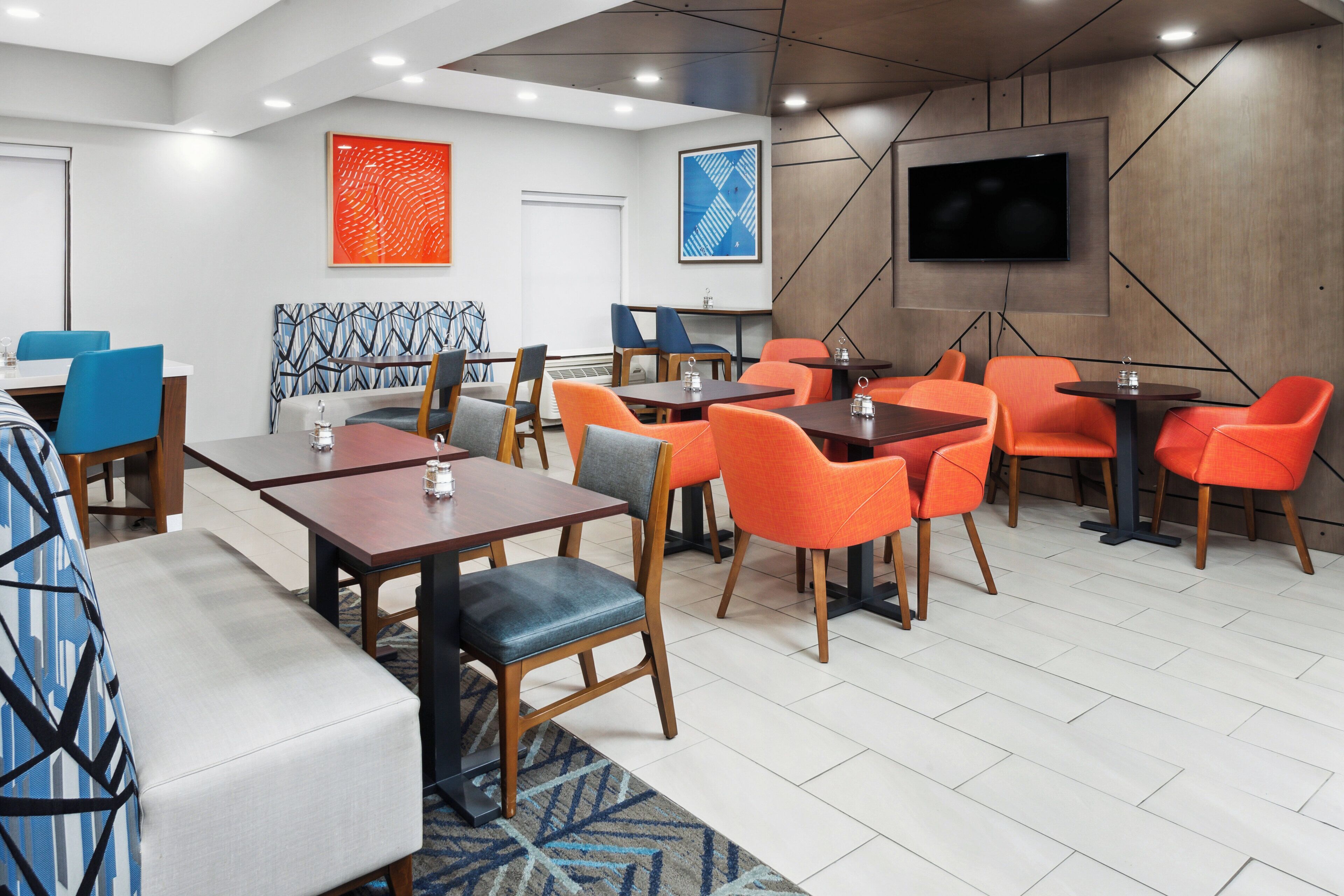En modern hotellobby med en blandning av olika sittplatser, inklusive bord och stolar, en stor platt-TV som är monterad på väggen och en mönstrad matta på golvet.
