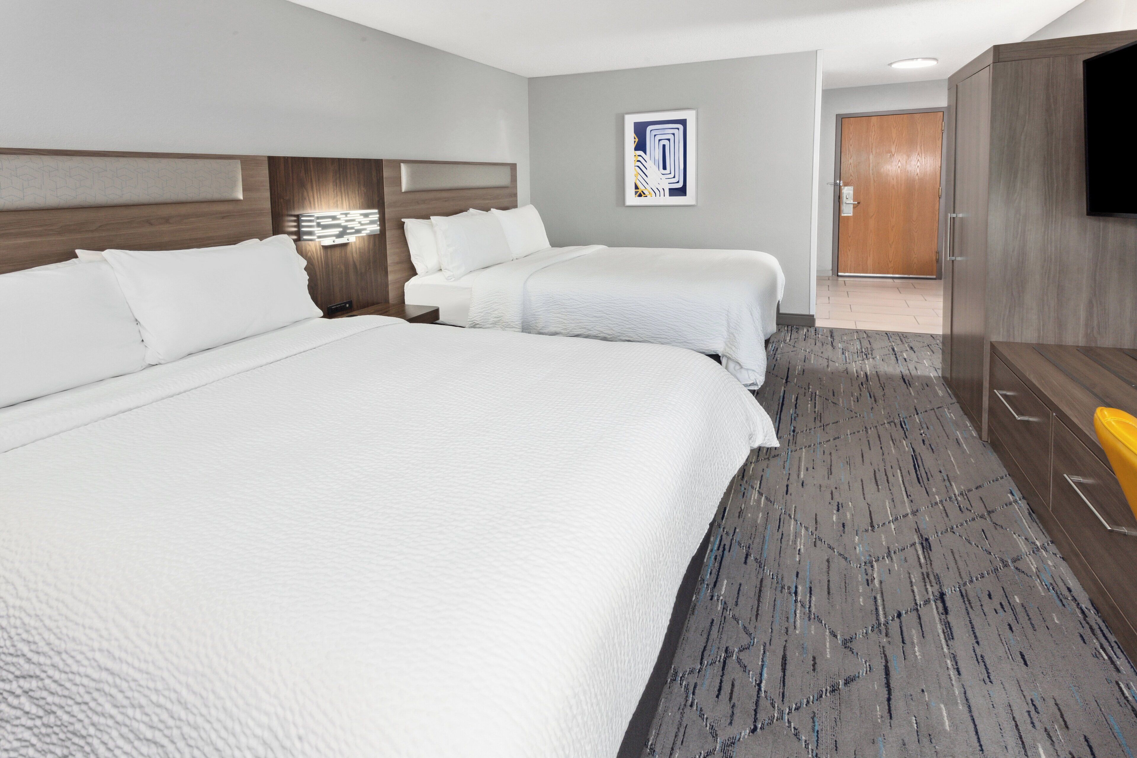 A hotel room with two beds, a wooden headboard, a gray carpet, a wooden wardrobe, and a flat-screen TV mounted on the wall.