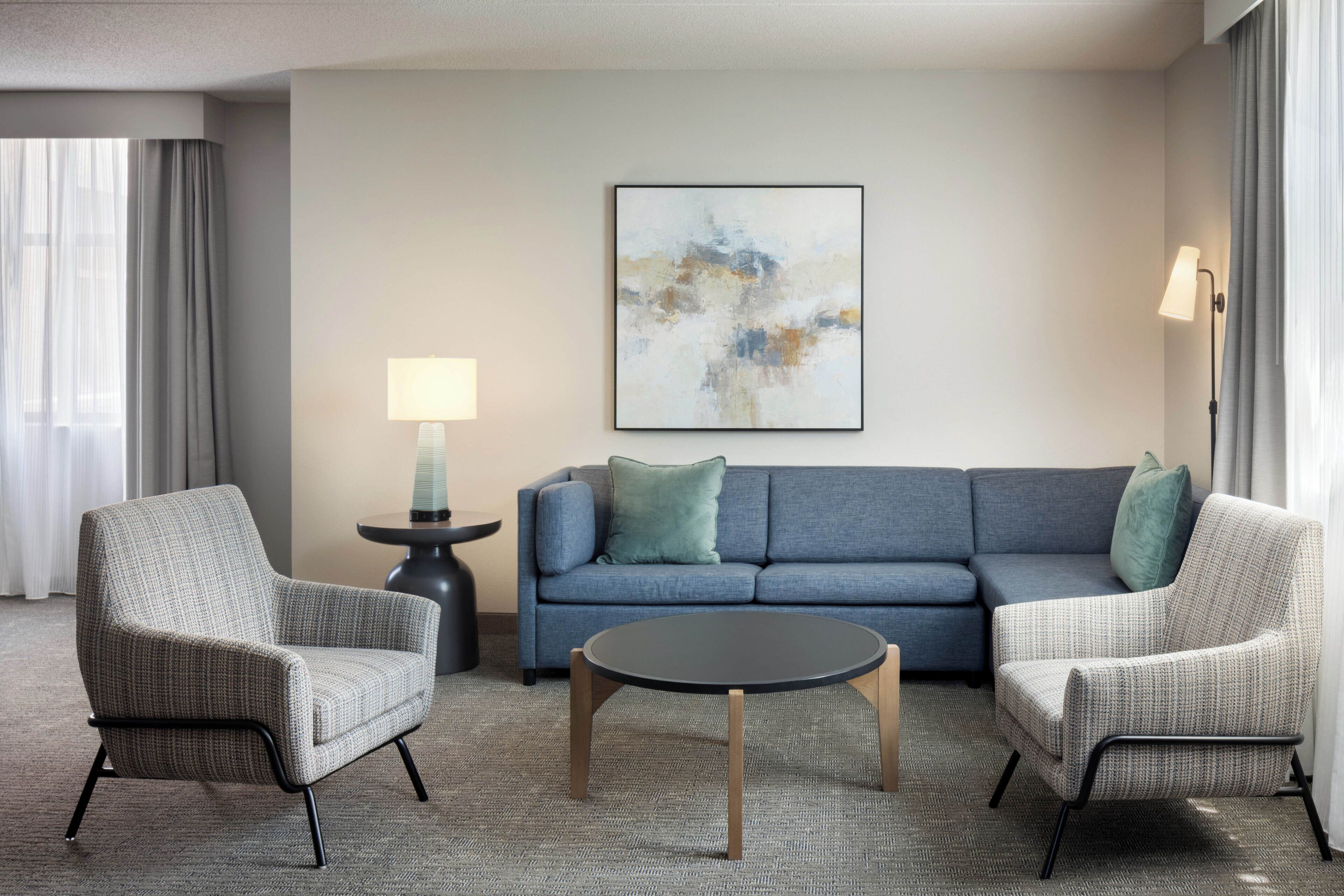A hotel room with a blue sofa, two armchairs, a coffee table, a side table with a lamp, and a painting on the wall.