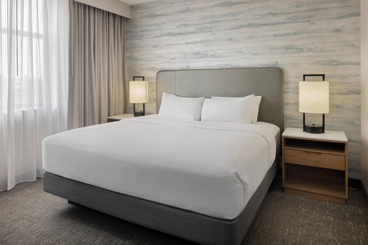 Ein Hotelzimmer mit einem großen Bett, zwei Nachttischen mit Lampen, einem Fenster mit durchsichtigen Vorhängen und einer Wand mit Marmoroptik.