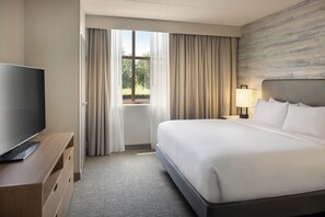 Habitación de hotel con una cama, un televisor sobre un soporte de madera, una mesita de noche con lámpara y vistas al exterior a través de una ventana con cortinas.