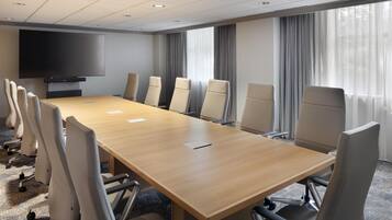 Un salón de conferencias con una larga mesa, sillas, un televisor de pantalla plana y ventanas grandes con cortinas.