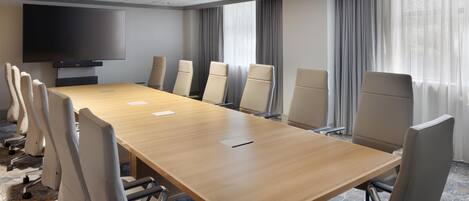 A conference room with a long table, chairs, a flat-screen TV, and large windows with curtains.