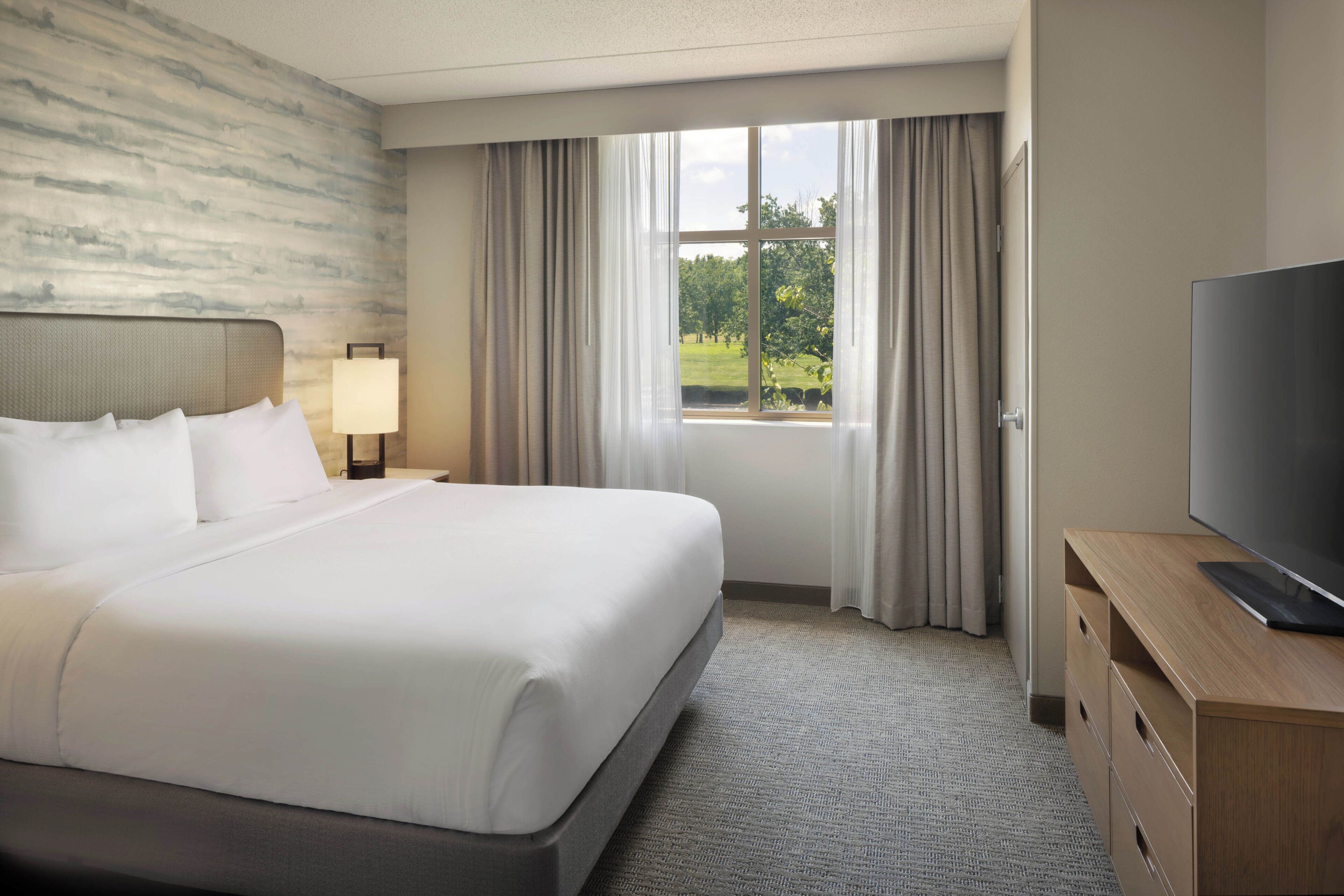A hotel room with a large bed, a television on a wooden dresser, and a window with curtains.