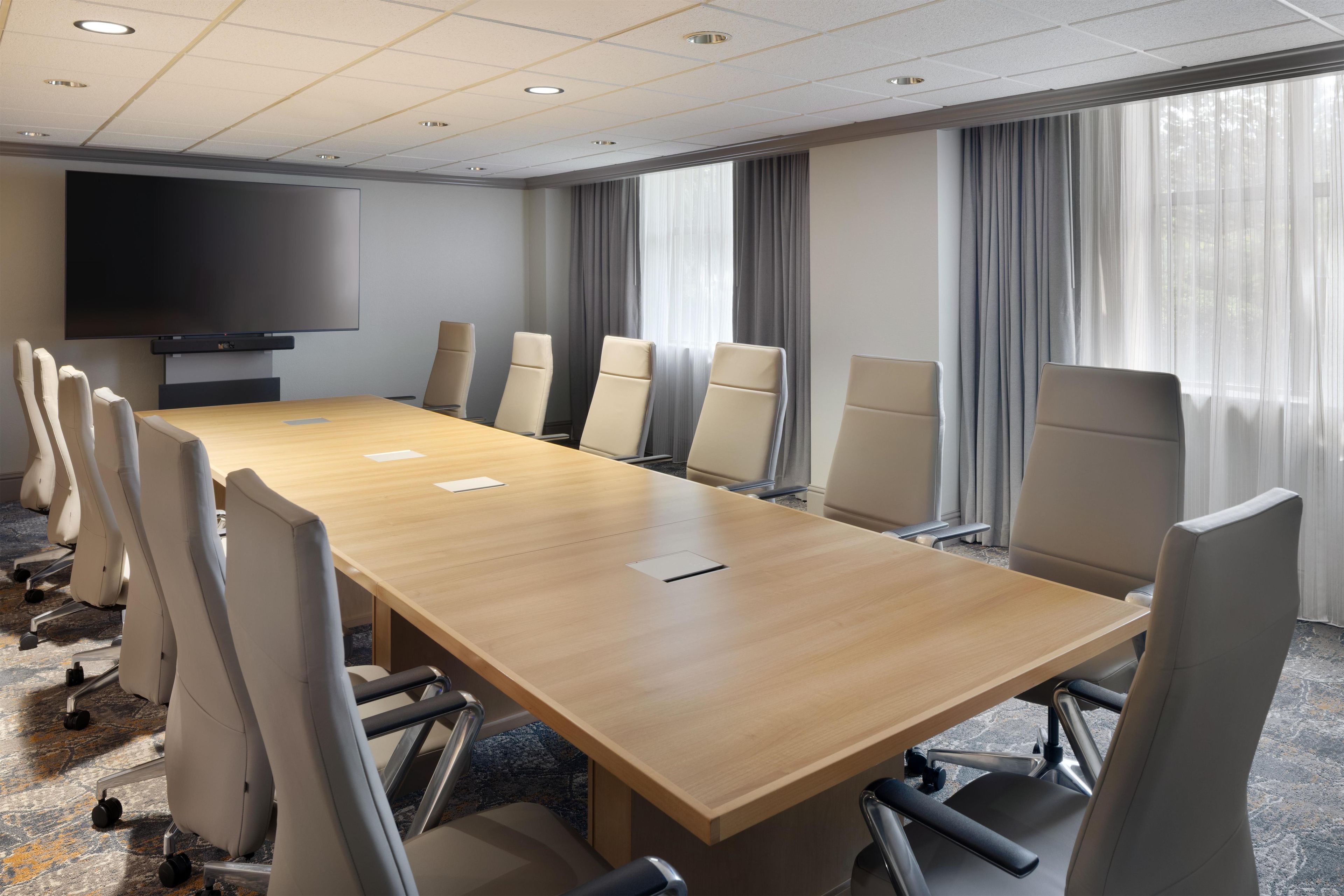 A conference room with a long table, chairs, a flat-screen TV, and large windows with curtains.