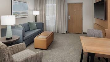 Habitación de hotel con sofá, mesa, televisión, puerta y ventana con cortinas.