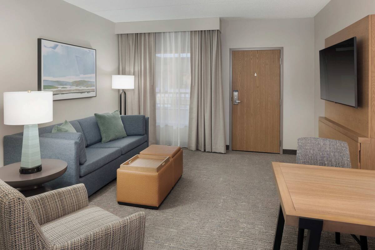 Ein Hotelzimmer mit Sofa, Hocker, Tisch, Lampe, Fernseher und Vorhängen.