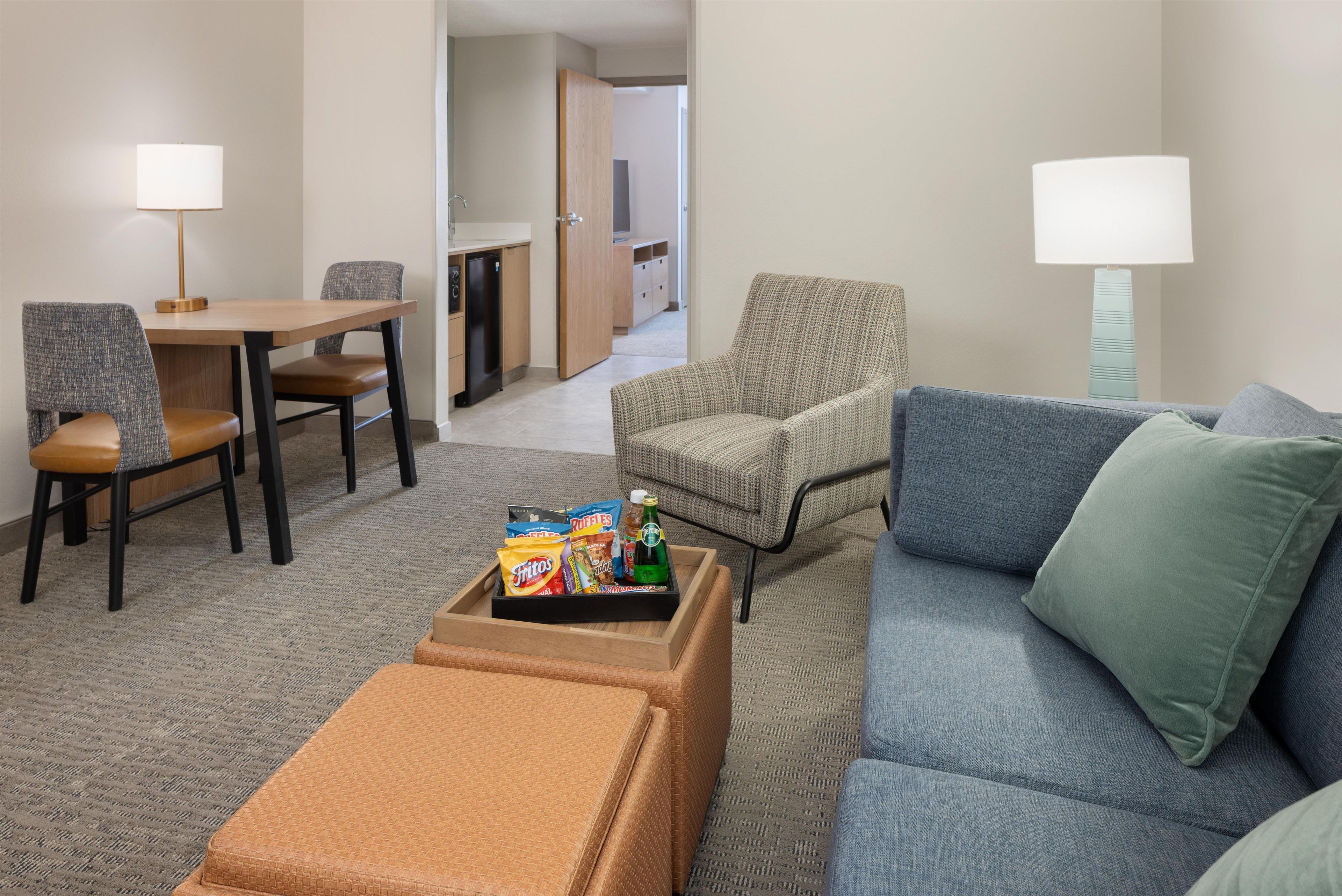 A hotel room with a dining area, a sofa, a chair, and a small table with snacks.