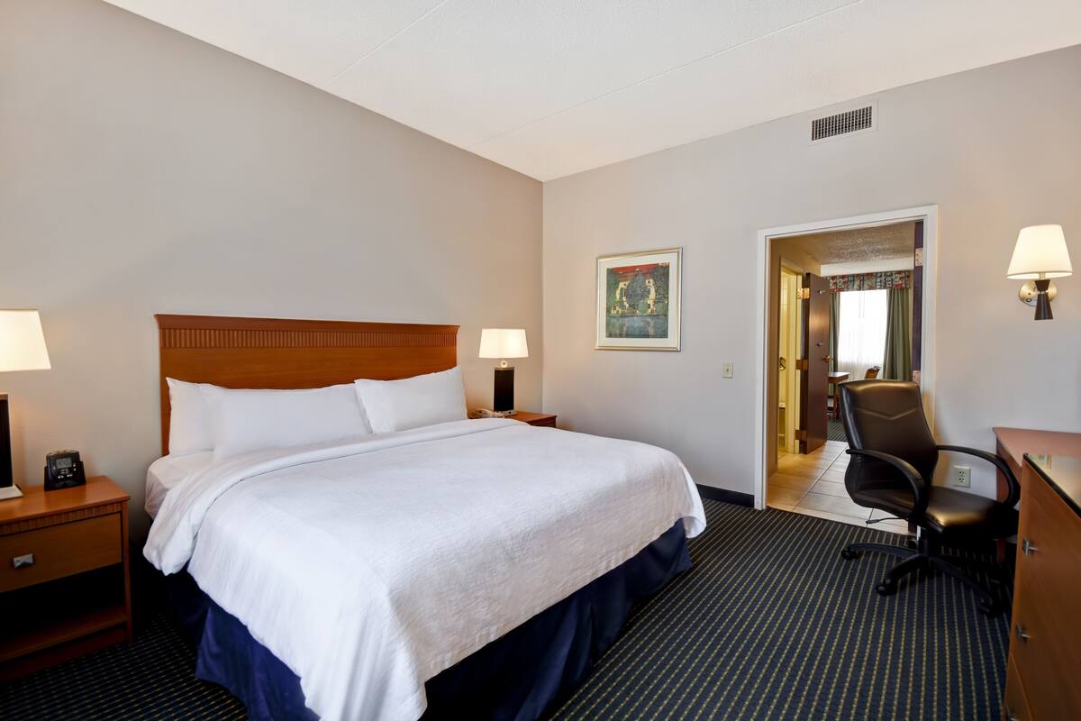 A hotel room with a large bed, bedside lamps, a desk with a chair, and a door leading to another room.